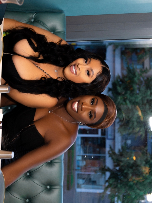 Two Women Smiling For A Photo While Seated At A Table In A Restaurant With Large Windows.